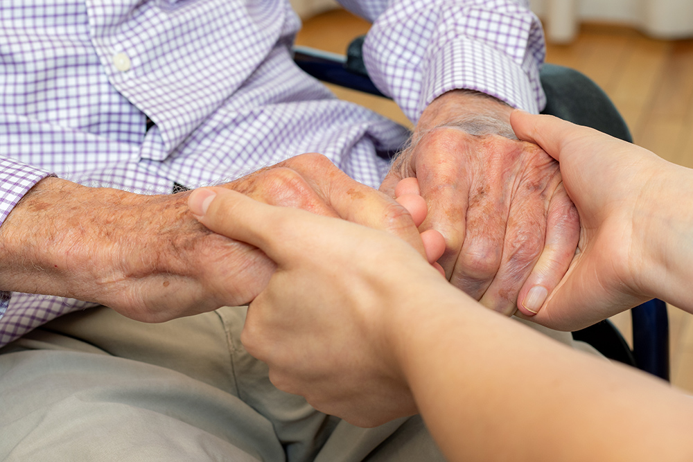 Close up detail of young female caregiver holding hands with old man in wheelchair.